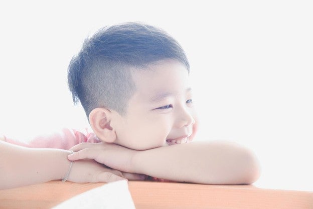 เด็กผมสีเข้มกำลังยิ้มและพักศีรษะบนแขน มองไปทางขวาโดยมีพื้นหลังสีขาวสว่าง