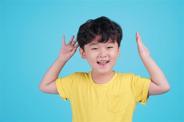  เด็กชาตัวเล็กทรงผมสไตล์เกาหลี สวมเสื้อยืดสีเหลืองยกมือขึ้นทั้งสองข้างบนพื้นหลังสีฟ้า